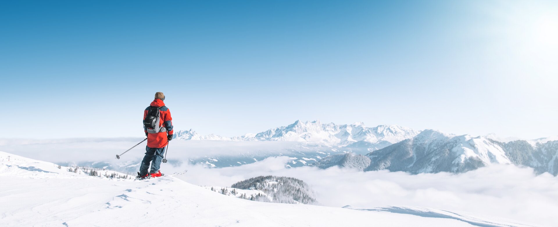 Skifahrer mit roter Skijacke steht  auf dem Schnee und Blickt die umliegenden Berge an