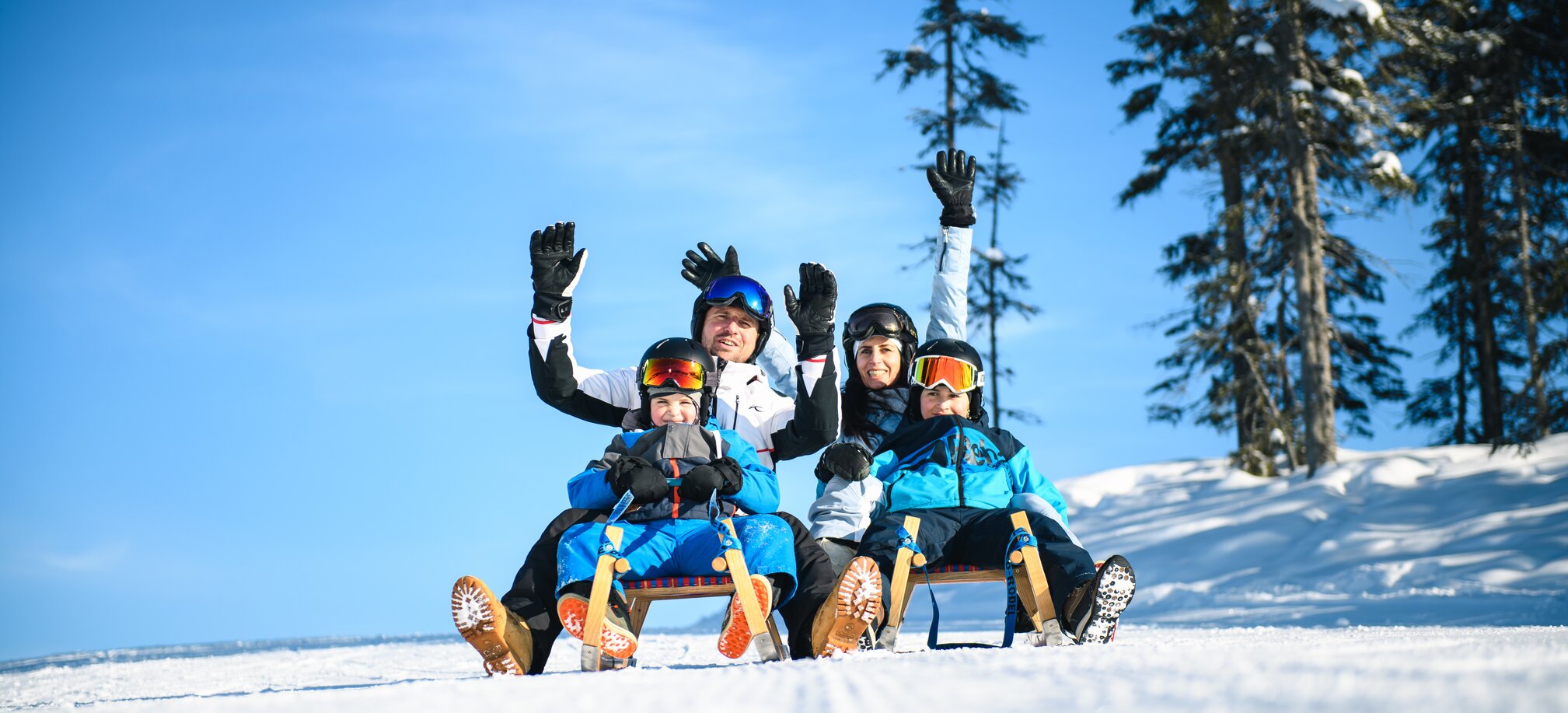 The father and mother each sit on a toboggan with a child in front of them and hold their hands up in joyful anticipation