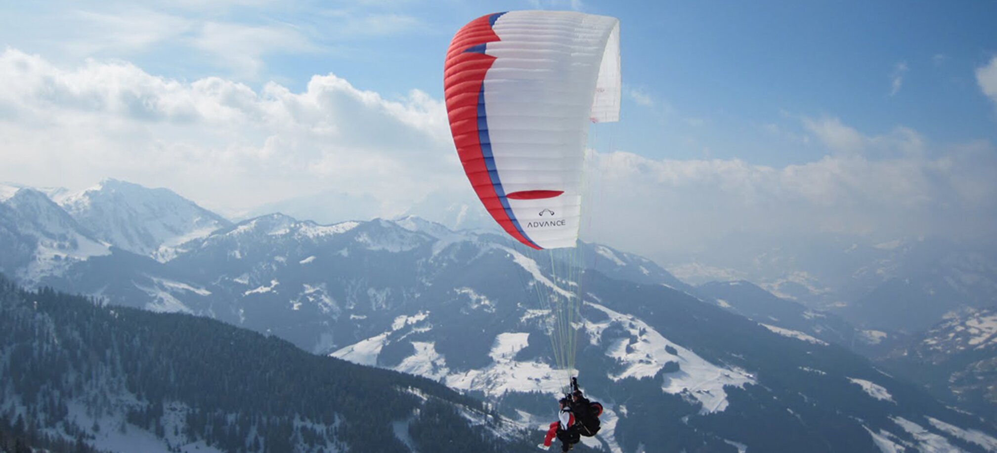 Paraglider with red, blue and white glider flies over mountains covered in forest and snowy pistes