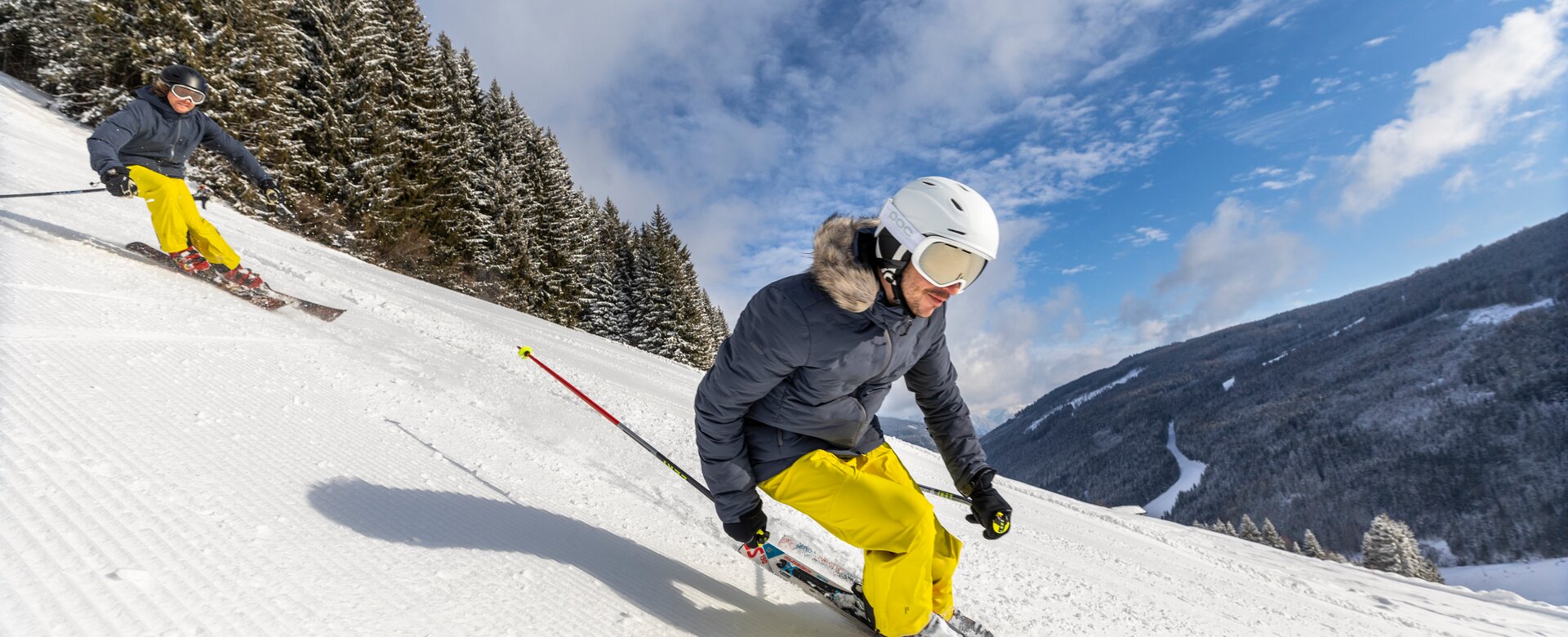 Zwei Skifahrer jeder mit gelber Hose und grauer Jacke legen sich in die Kurve um die Piste hinunterzufahren