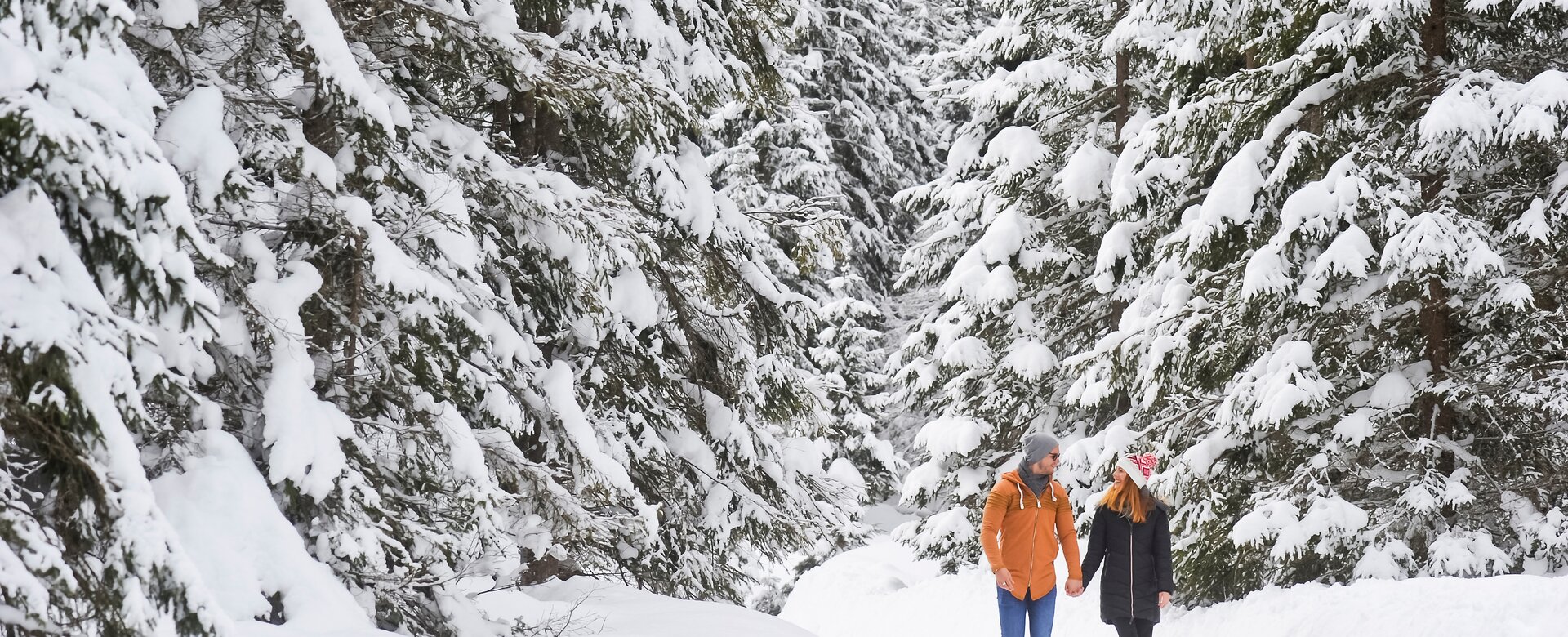 Ein Paar geht Händchen haltend auf einem Weg aus Schnee zwischen verschneiten Bäumen und lächelt sich an | © Huber Hans Photos