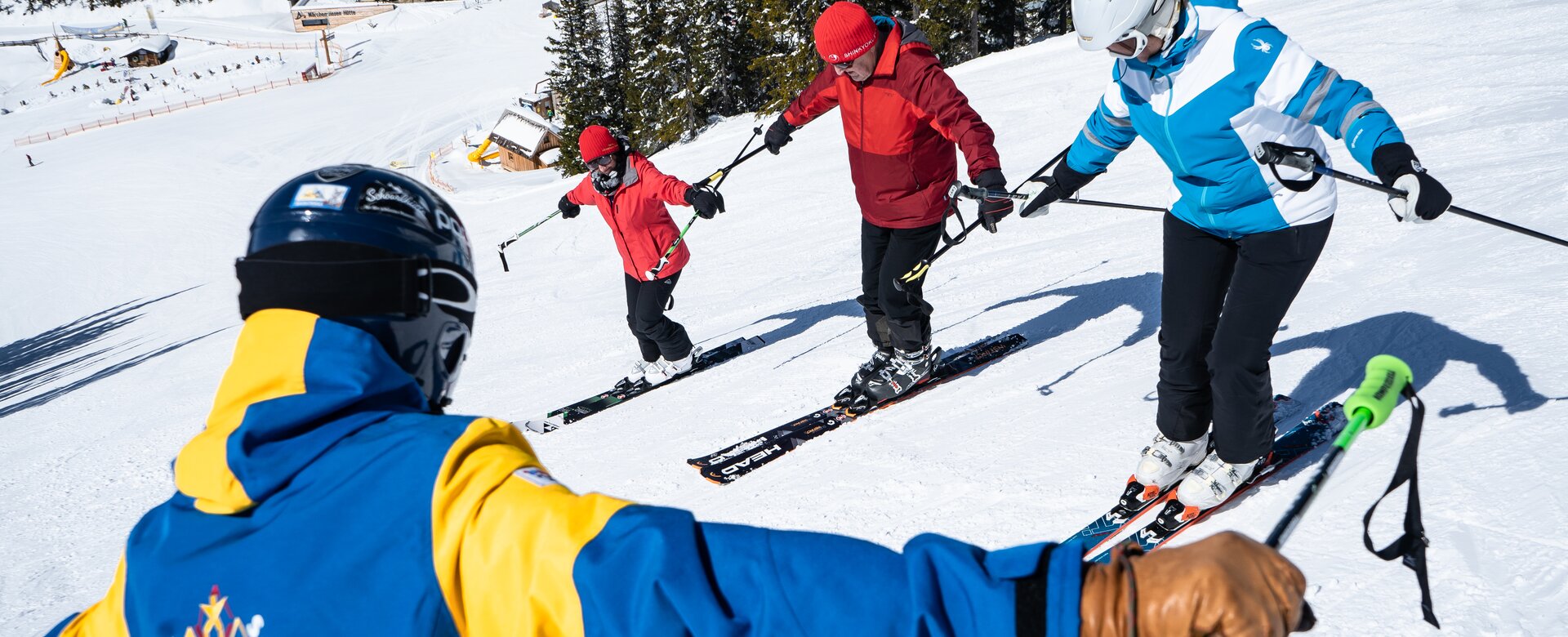 A ski instructor in a blue and yellow ski suit shows three students a balancing exercise with outstretched arms.